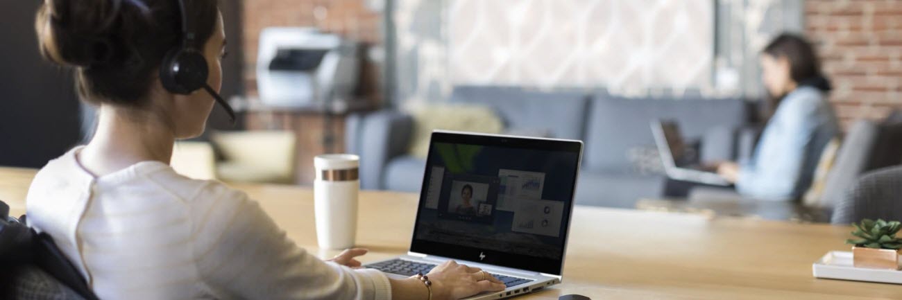 A woman at a large wooden desk, wearing a headset, video-conferencing on the open laptop in front of her