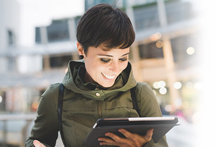 A short-haired woman looking at her tablet while she is outside a building complex