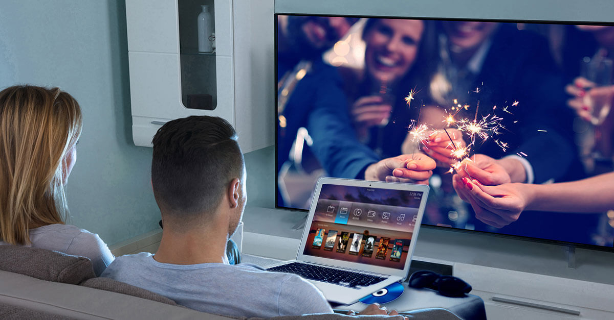 A man and woman sitting on a couch with their open laptop that has a CD coming out, all in front of a projector screen showing a photo of a group of friends putting together burning sparkler fireworks that they're holding
