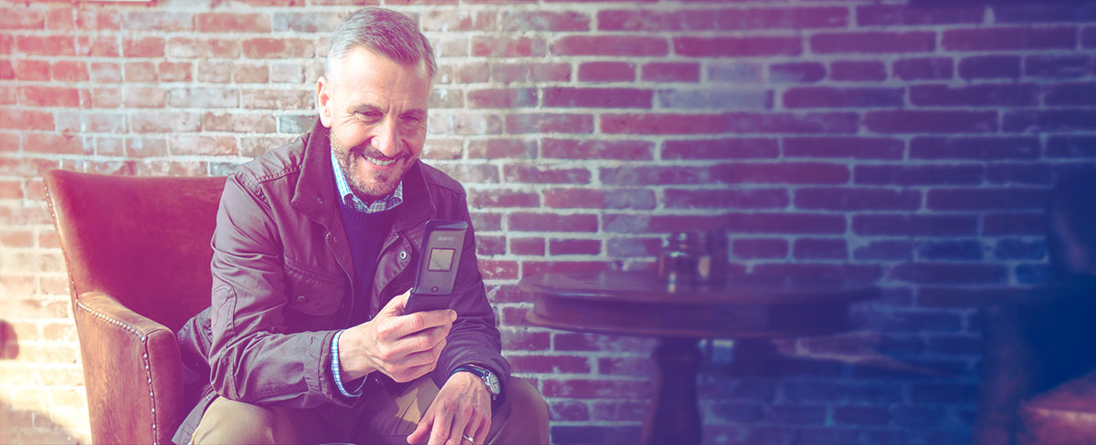 An older gentleman in a brick-walled coffee shot smiling at his open Alcatel A405 cell phone