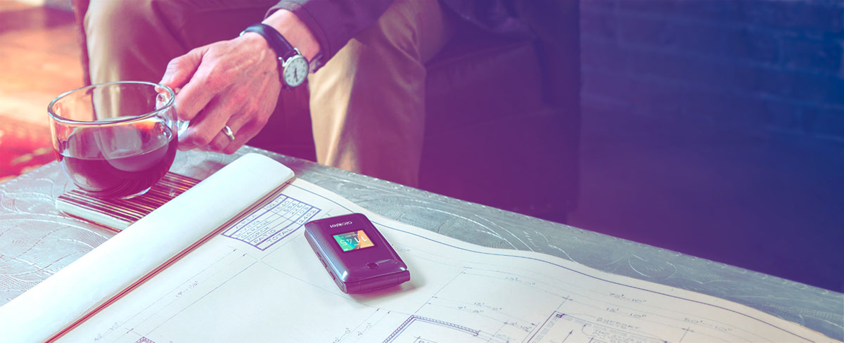 The Alcatel A405 cell phone laying flat on top of blueprints on a metal table with a hand grabbing for a cup of coffee that's next to the phone