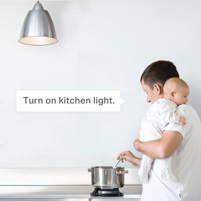 voice command bubble for 'turn on kitchen light' in front of a man holding his baby over his left shoulder while he cooks something in a pot on a stove