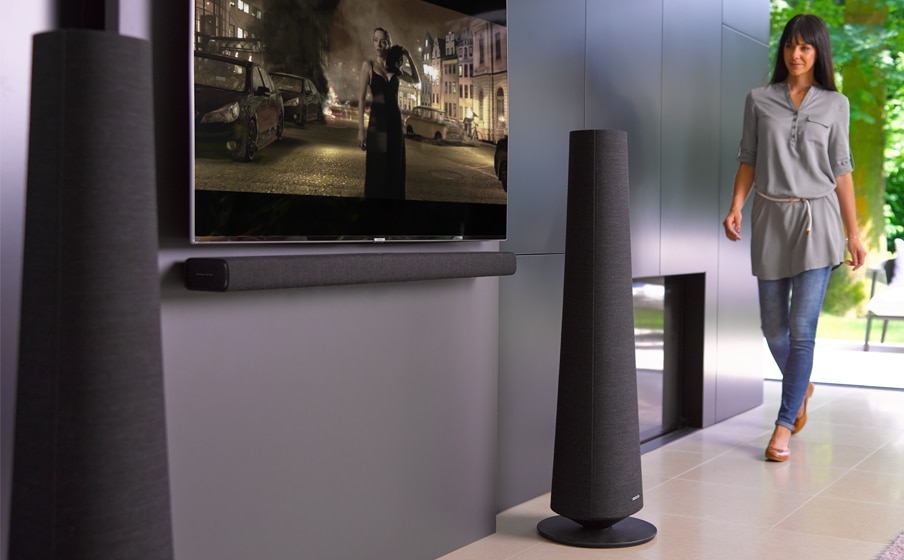 A wowman walking towards her TV and harman kardon citation speakers on the floor with a mounted TV in between