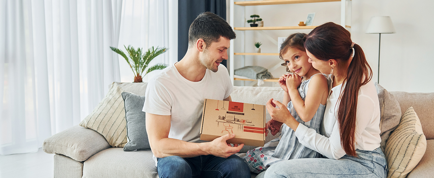 Family scene on couch: man, woman, and young girl opening a cardboard box together, surrounded by living room decor including a plant.