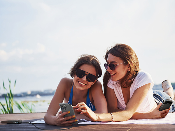Two people lying on a surface near water, looking at a smartphone together. Sunny outdoor setting with vegetation in background.