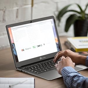 Laptop on desk showing search engine homepage. Person's hands visible typing on keyboard, with plant in background.