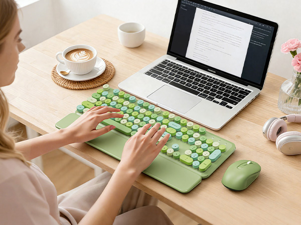 Person typing on green keyboard with wrist rest and mouse beside a laptop on desk