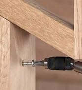 Close-up of a power drill driving a screw into light-colored wood, likely part of furniture assembly or construction.