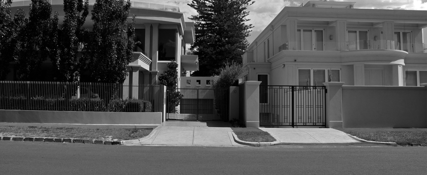 Black and white image of a gated property entrance. Shows a large house behind iron fencing, with trees and a sidewalk visible in the foreground.