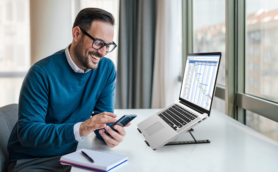 Person at desk working with mobile phone and laptop on elevated stand near window in office setting.