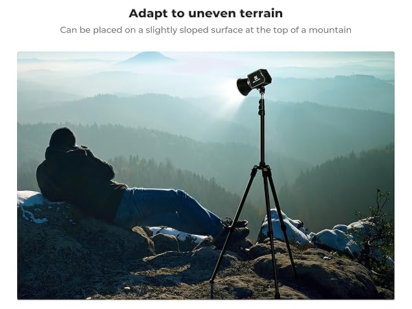 Camera tripod on rocky mountain peak, demonstrating adaptability to uneven terrain. Person and gear visible, overlooking misty forested valleys in background.