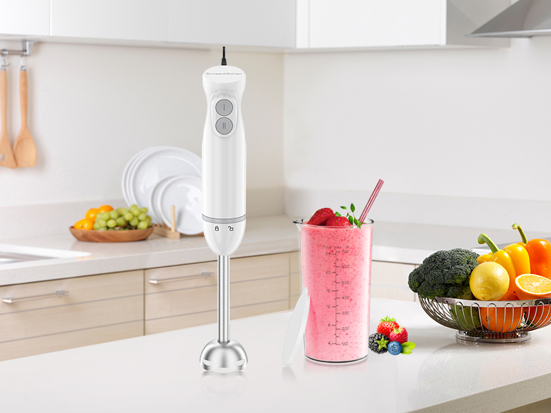 White kitchen counter setup with immersion blender and measuring cup. Fresh fruits and smoothie ingredients visible alongside blending equipment.