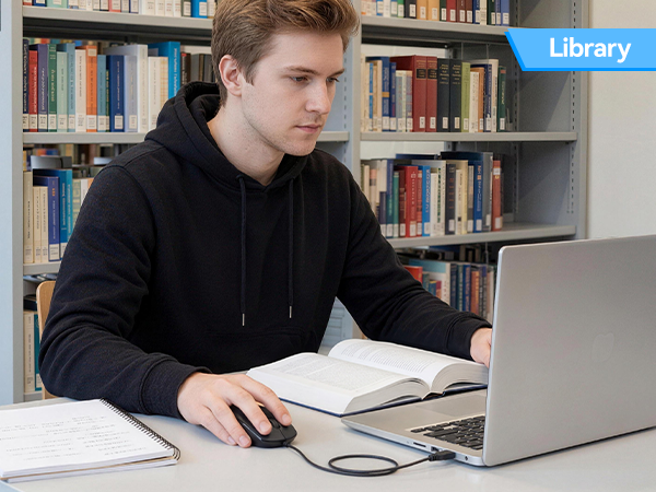 Text reads 'Library'. Young man in black hoodie studying at table with open notebook and silver laptop, bookshelves visible in background.