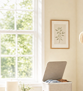Interior room setting with beige/tan colored wall, window with natural light, and framed artwork mounted on wall above a light-colored chair.