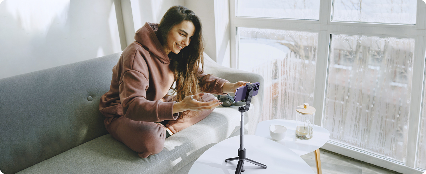 Smartphone tripod in use. Woman on couch holds phone mounted on adjustable black stand. Flexible legs allow positioning on uneven surfaces.