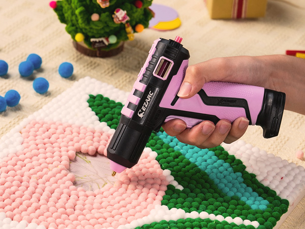 Pink and black cordless hot glue gun being used on colorful textile crafts, with decorative elements in background showing practical application.