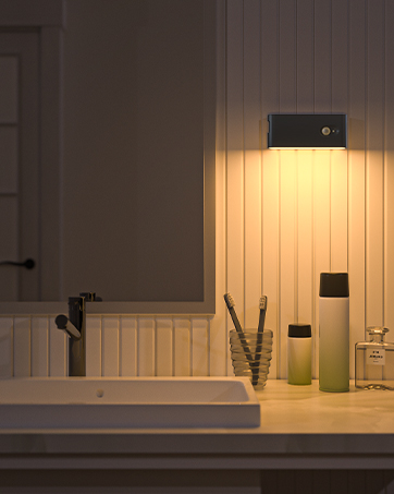 Bathroom vanity with illuminated mirror, white sink, and wooden cabinetry. Warm lighting highlights toiletries on countertop.