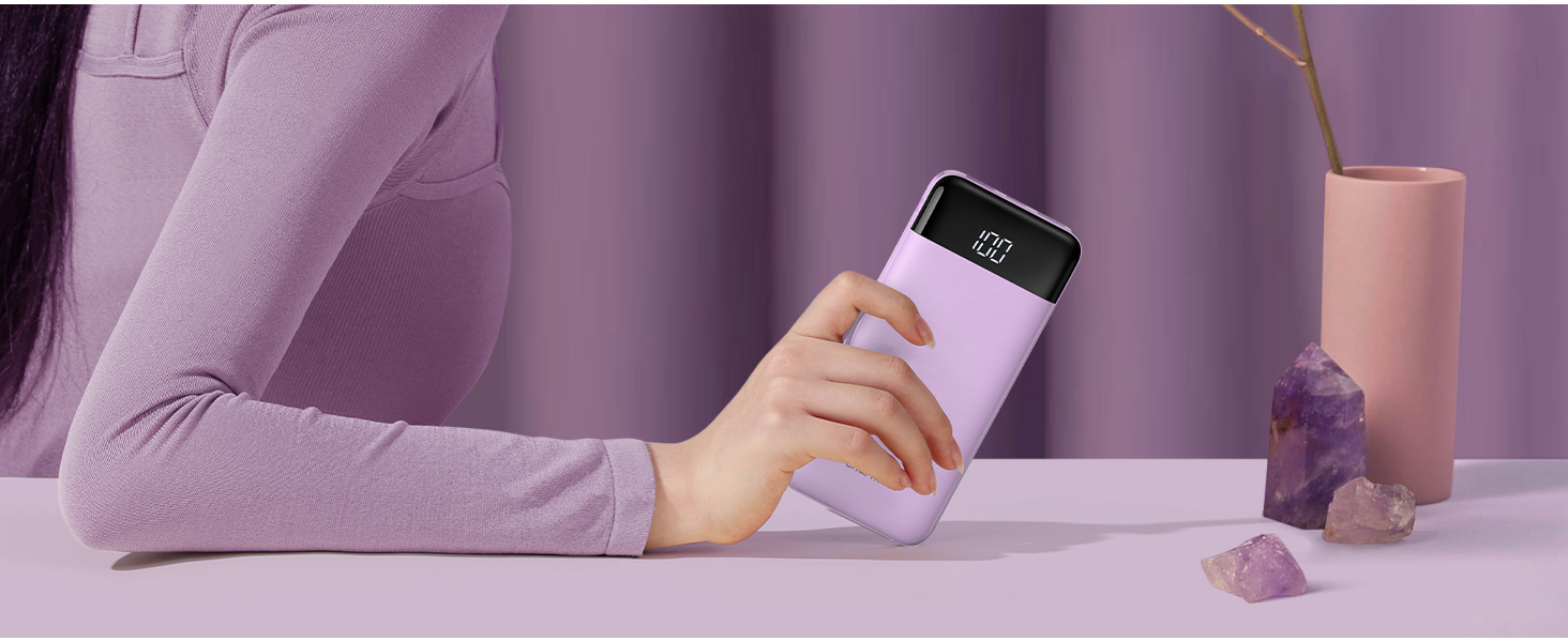 Hands holding a lilac-colored cylindrical device with a digital display against a purple background. A vase and decorative object are visible.