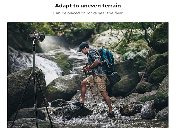 Tripod with flexible legs on rocky terrain near a stream. Hiker with camera and backpack navigates rocks, demonstrating tripod's adaptability to uneven surfaces.
