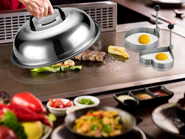 Stainless steel dome lid covering food on cooking surface, with various condiments and ingredients arranged nearby including peppers and sauces.
