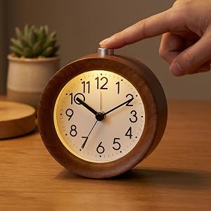 Wooden round analog alarm clock with brown frame, white face, and black numerals 1-12 displayed on wooden surface with small potted succulent plant beside it.