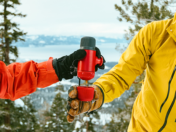 Red insulated beverage container being shared between two people wearing winter gloves and colorful jackets against a snowy mountain backdrop.
