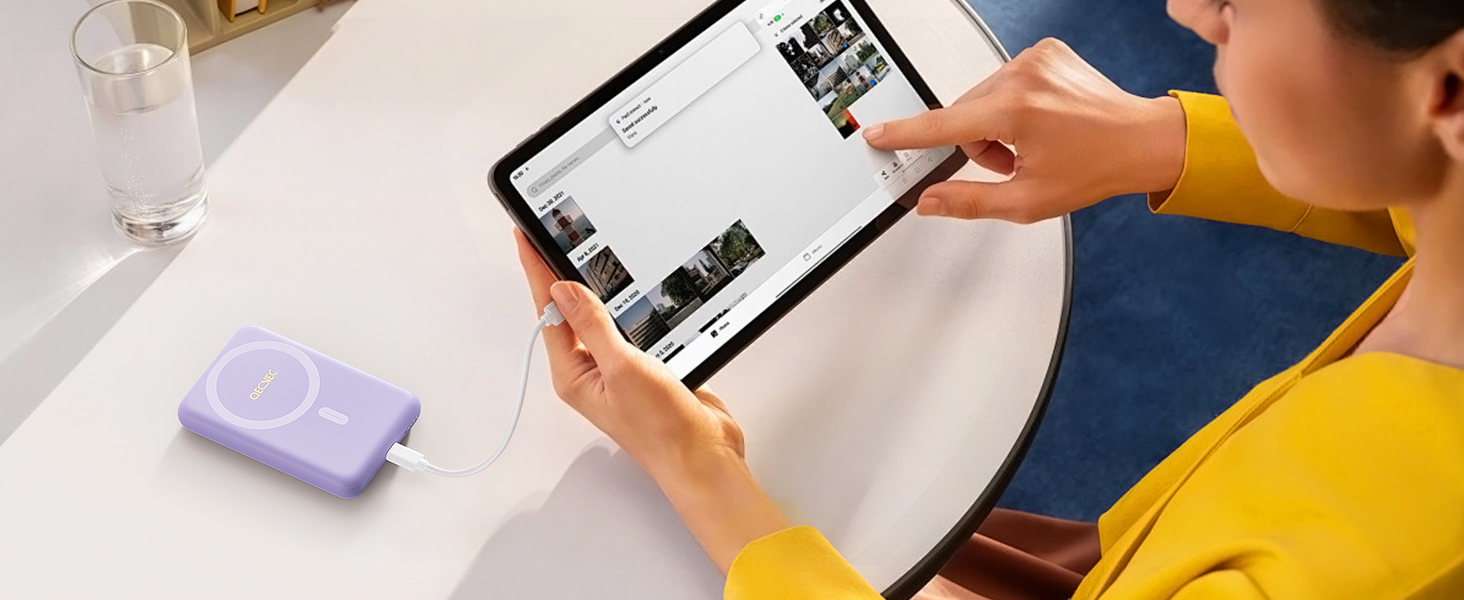 Hands holding a tablet displaying a photo gallery app. A small purple rectangular device with rounded corners sits on the table nearby.