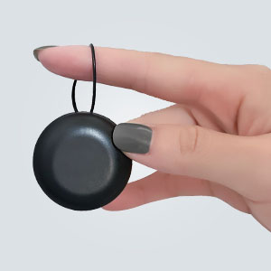 Small round black device held between thumb and forefinger. Thin cord attached, suggesting it's a wearable or portable electronic accessory.