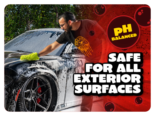 Smiling detailer sprays pink cherry foam from a foam gun across a dark SUV in bright sunlight