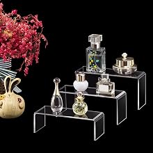 Clear acrylic display stands arranged in ascending steps, showcasing perfume bottles and decorative items against a black background with pink flowers.