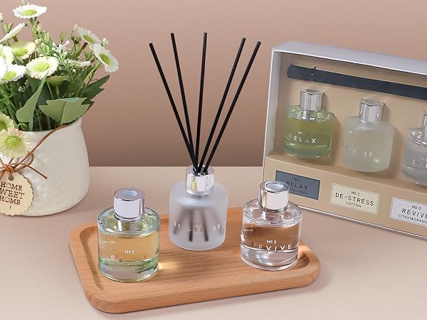 Home fragrance display on wooden tray featuring reed diffuser, scented candles, and decorative packaging, alongside small potted white flowers in burlap-wrapped container.