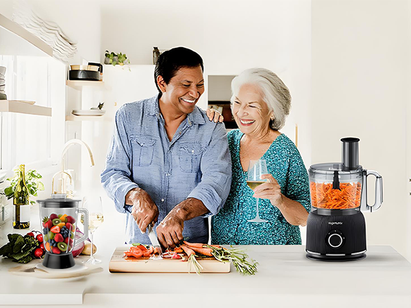 Kitchen scene showing food preparation with fresh vegetables and an electric food processor on a white countertop.