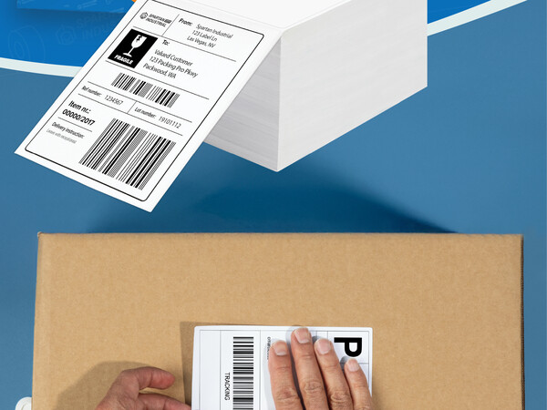 A man applies a shipping tag to a cardboard box next to a stack of fanfold thermal labels
