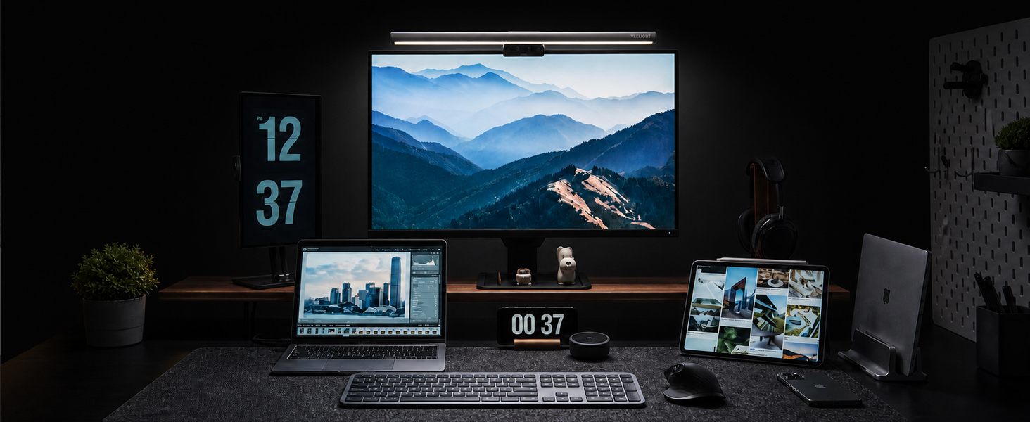 Modern desk setup with dual monitors displaying mountain landscape and time displays, laptop, desktop computer tower, and office accessories on dark surface against black wall.