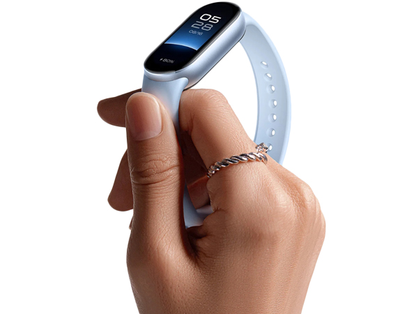 White fitness tracker with oval display held between thumb and forefinger. Device shows time on screen and has a white silicone strap.