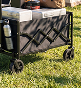 Black collapsible utility cart with X-frame design and wheels on grass outdoors, featuring storage compartment and handle.