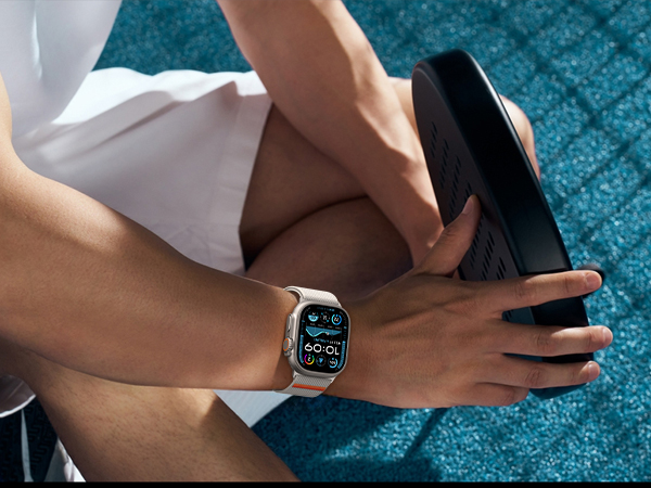 Smartwatch with a square face and light-colored band on a person's wrist. The watch face displays colorful app icons. The wearer is holding a dark object, possibly exercise equipment.