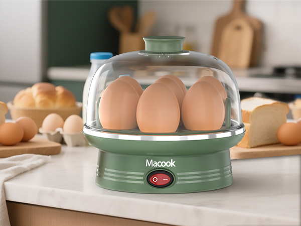Macook egg cooker with clear dome lid, showcasing six brown eggs inside. Green base with power switch, sitting on kitchen counter with bread and additional eggs nearby.