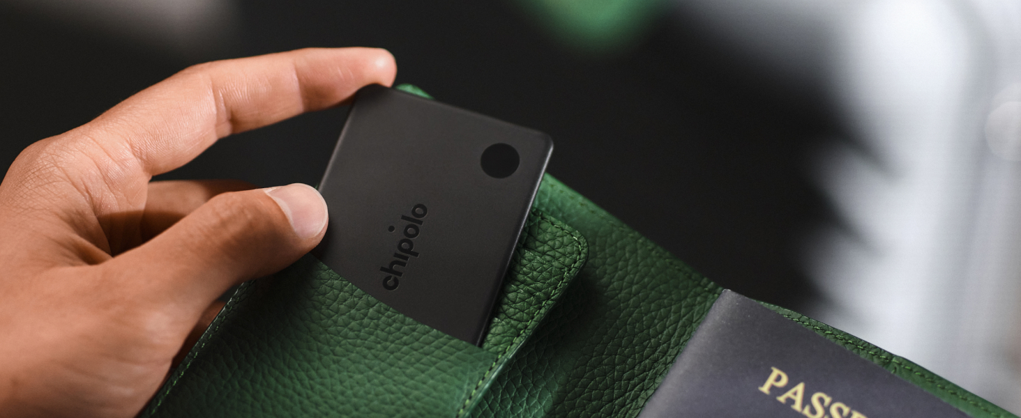 Close-up of hand holding black rectangular measuring device with green fabric strap. Detail view shows compact sensor tool with carrying attachment in outdoor setting.