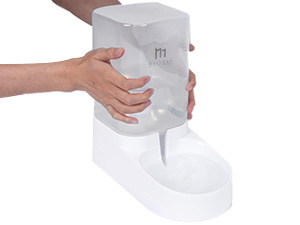 Hands holding a large, cylindrical white dispenser with a curved base. The dispenser has a visible spout and appears to be made of plastic.