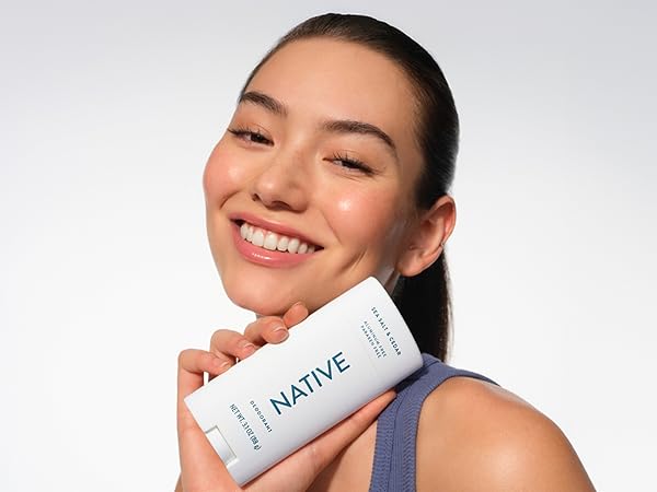 Smiling woman with fresh skin holds Native Sea Salt & Cedar deodorant, looking confident.