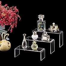 Clear acrylic display stands arranged in ascending steps, showcasing perfume bottles and decorative items against a black background with pink flowers.