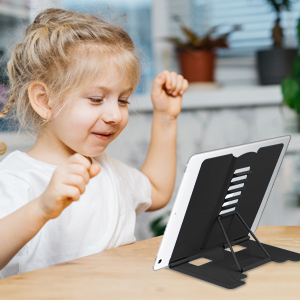 Child using a tablet device propped up on a black adjustable stand on a table, appearing engaged and happy.