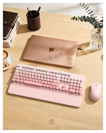Pink typewriter-style keyboard and mouse with wrist rest on a wooden desk beside a laptop and decor