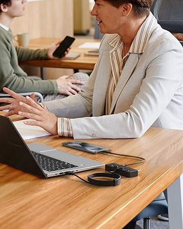 Person in light-colored blazer using laptop at wooden desk. Small round device visible near laptop, likely an audio accessory.