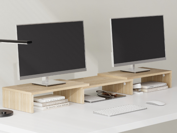Light wooden monitor riser shelf with multiple compartments spanning across two desktop monitors, providing storage space underneath for electronics and accessories.