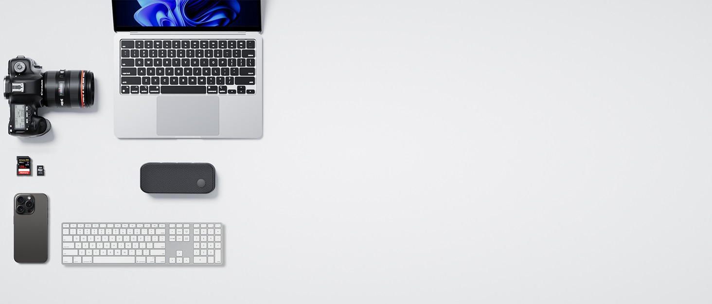 Overhead flat lay of computer workspace showing MacBook laptop, smartphone, wireless keyboard, mouse, and various tech accessories on white surface.