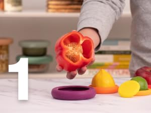 A red pepper is held over a silicone food saver with a lemon saver and lime saver on the counter
