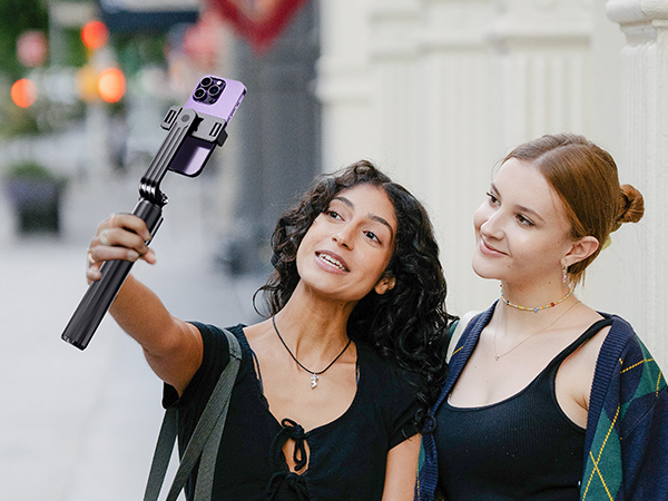 Extendable selfie stick with smartphone grip being used outdoors. Black telescoping pole with adjustable phone mount, demonstrated in use for taking photos.