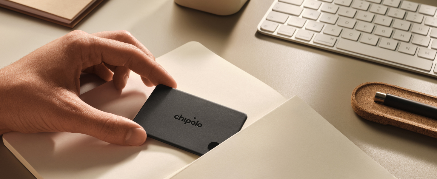 Hand holding a small dark rectangular wallet or card holder on a desk with keyboard and wooden accessories visible.